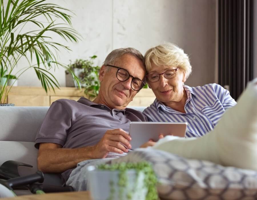 A man with a cast on his foot hugging a woman on a couch, both looking at a device