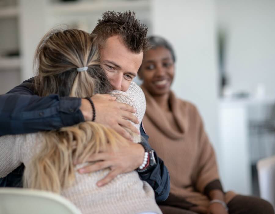 A man hugging a woman while another woman looks on, smiling, in the background