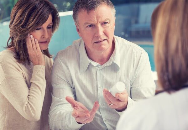 A man holding a pill bottle, and a woman looking upset, talking to a doctor